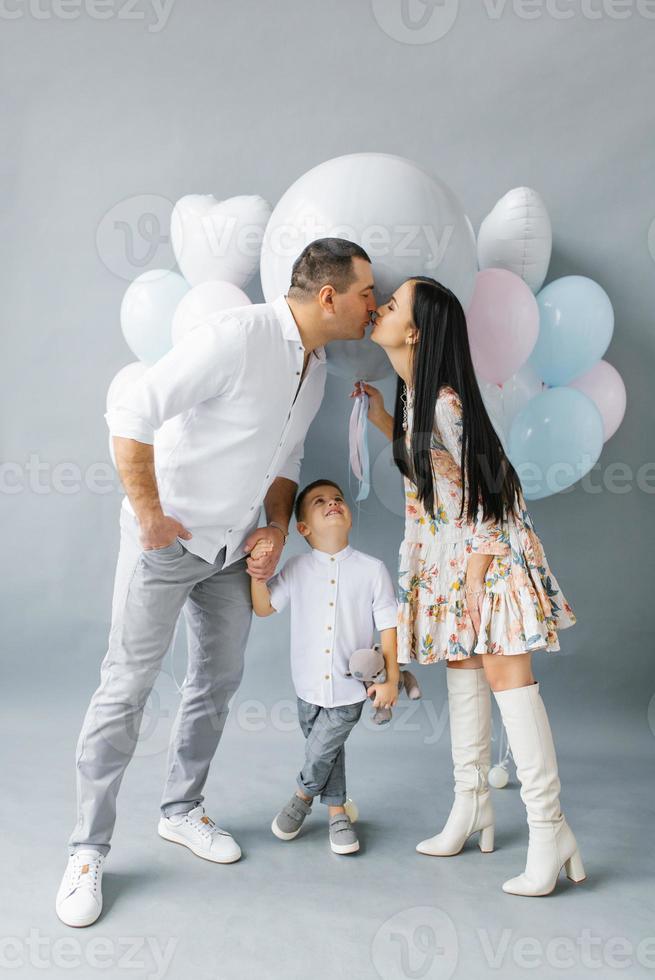 A young stylish family waiting for their second child is kissing against the background of a pop ball to determine the gender of the future baby. Parents with their eldest son on a gray background photo