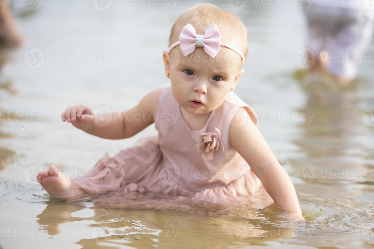 A beautiful little girl sits on the river bank and plays with sand and water. Child and nature. photo