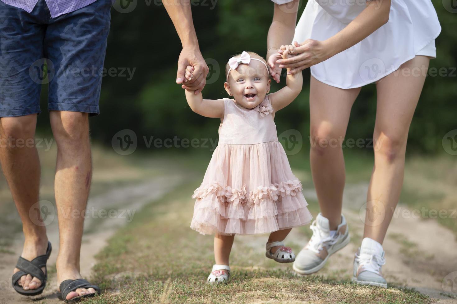Happy one-year-old girl is being held by dad and mom. Little child walking with parents. A cute baby learns to walk with the help of her parents. photo