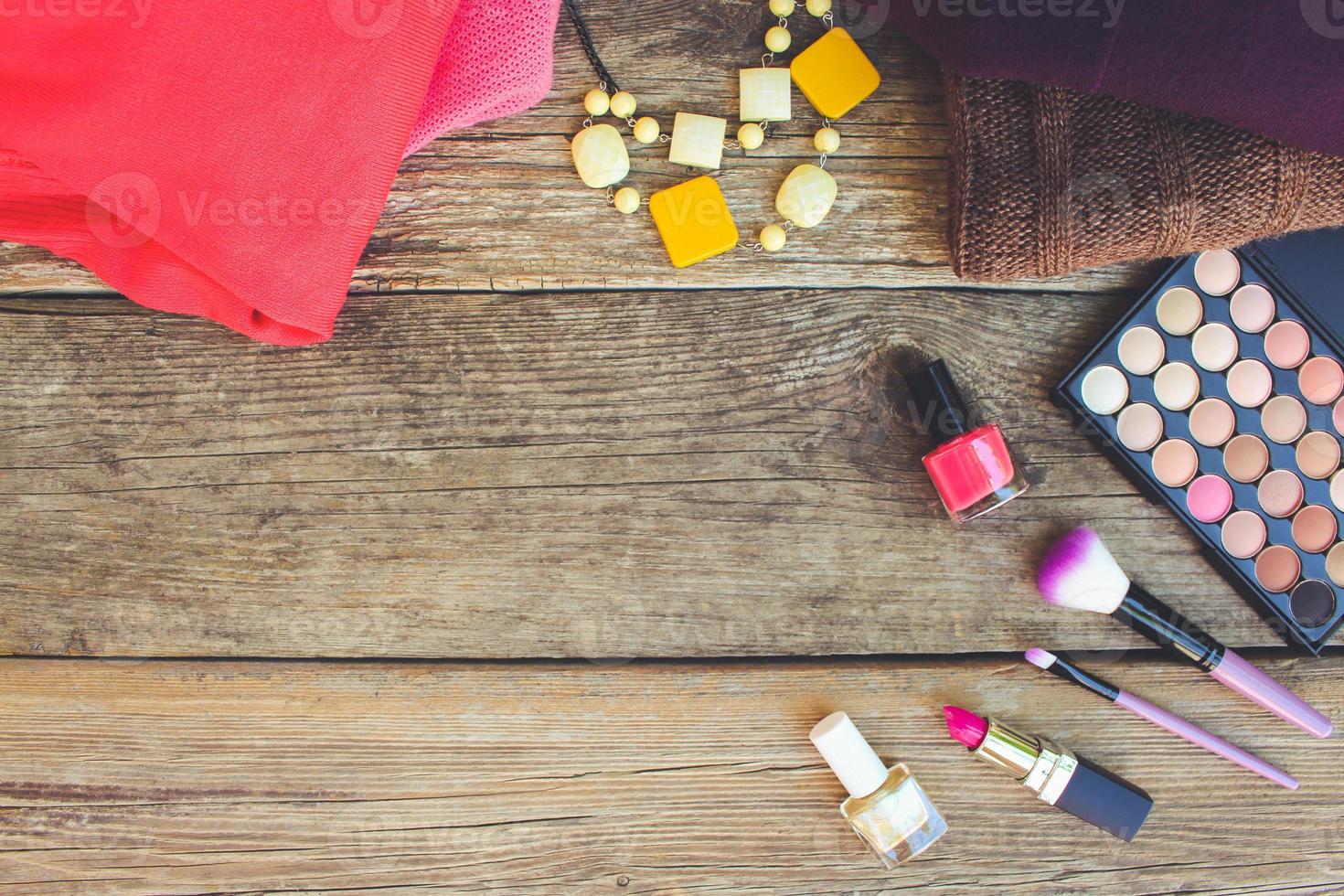 Women's clothing and cosmetics sweaters, lipstick, nail polish, necklaces, eye shadow, brushes on wooden background. Top view. Toned image. photo