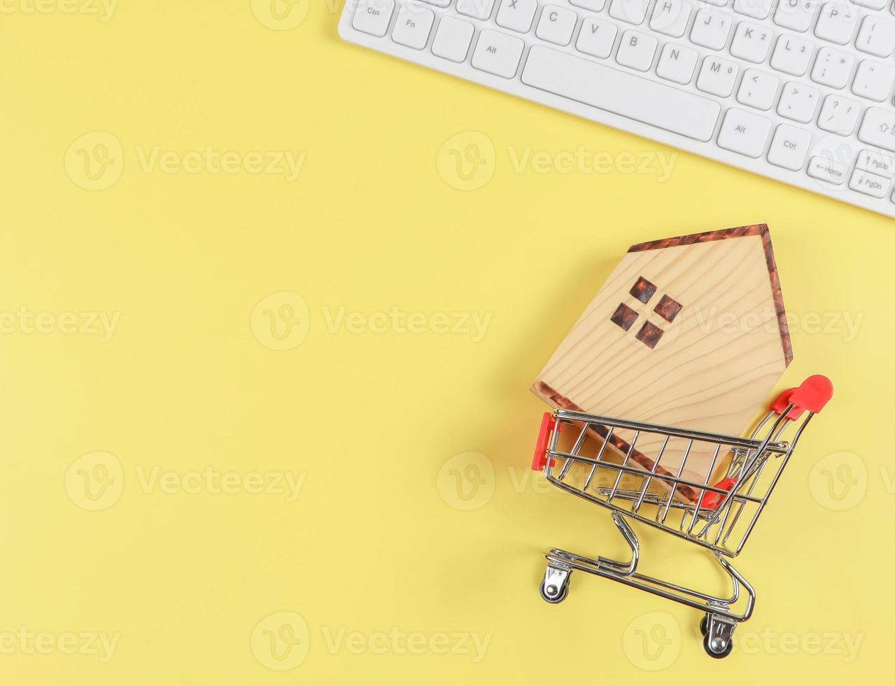 flat layout of wooden house model in shopping trolley and  and computer keyboard  on yellow background with copy space, home purchase concept. photo