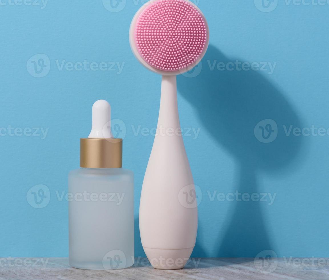 White glass bottle with a pipette and a plastic facial brush on a blue background photo