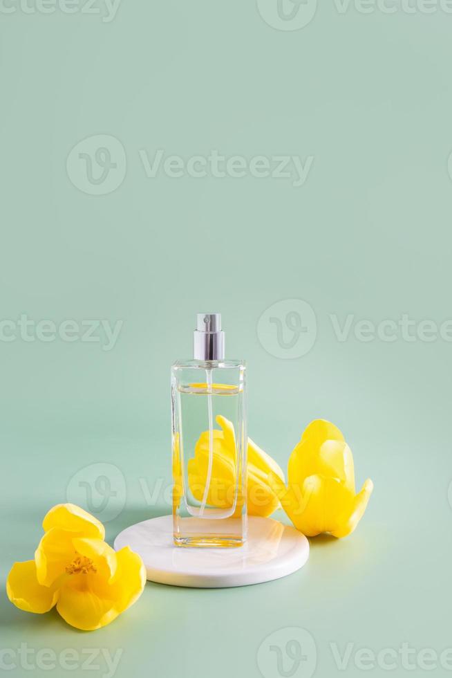a transparent bottle of cosmetic or spray stands on a white round podium with yellow tulips. vertical view. a copy space. green background. photo