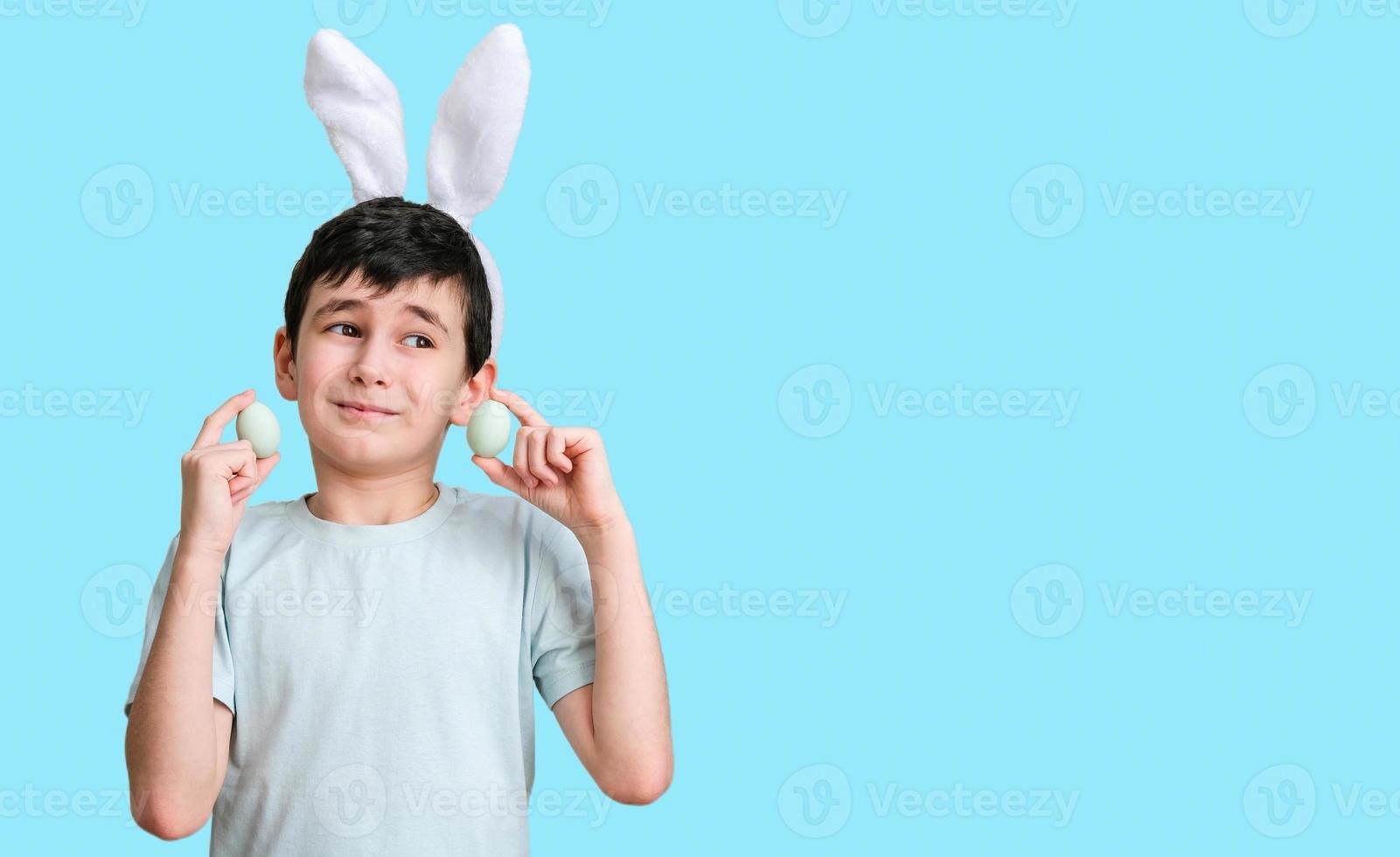 Smiling boy wearing bunny ears holding Easter eggs on a blue background with space for text. Easter banner photo