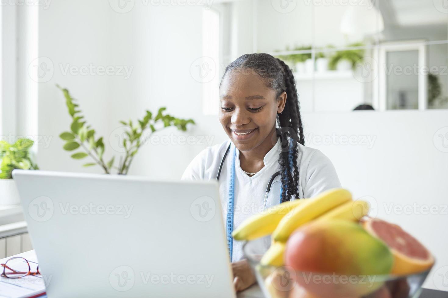 Portrait of young smiling female nutritionist in the consultation room. Nutritionist desk with healthy fruit, juice and measuring tape. Dietitian working on diet plan. photo