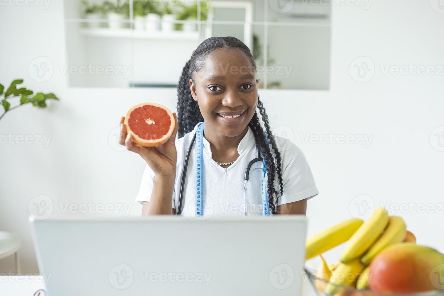 Portrait of young smiling female nutritionist in the consultation room. Nutritionist desk with healthy fruit, juice and measuring tape. Dietitian working on diet plan. photo