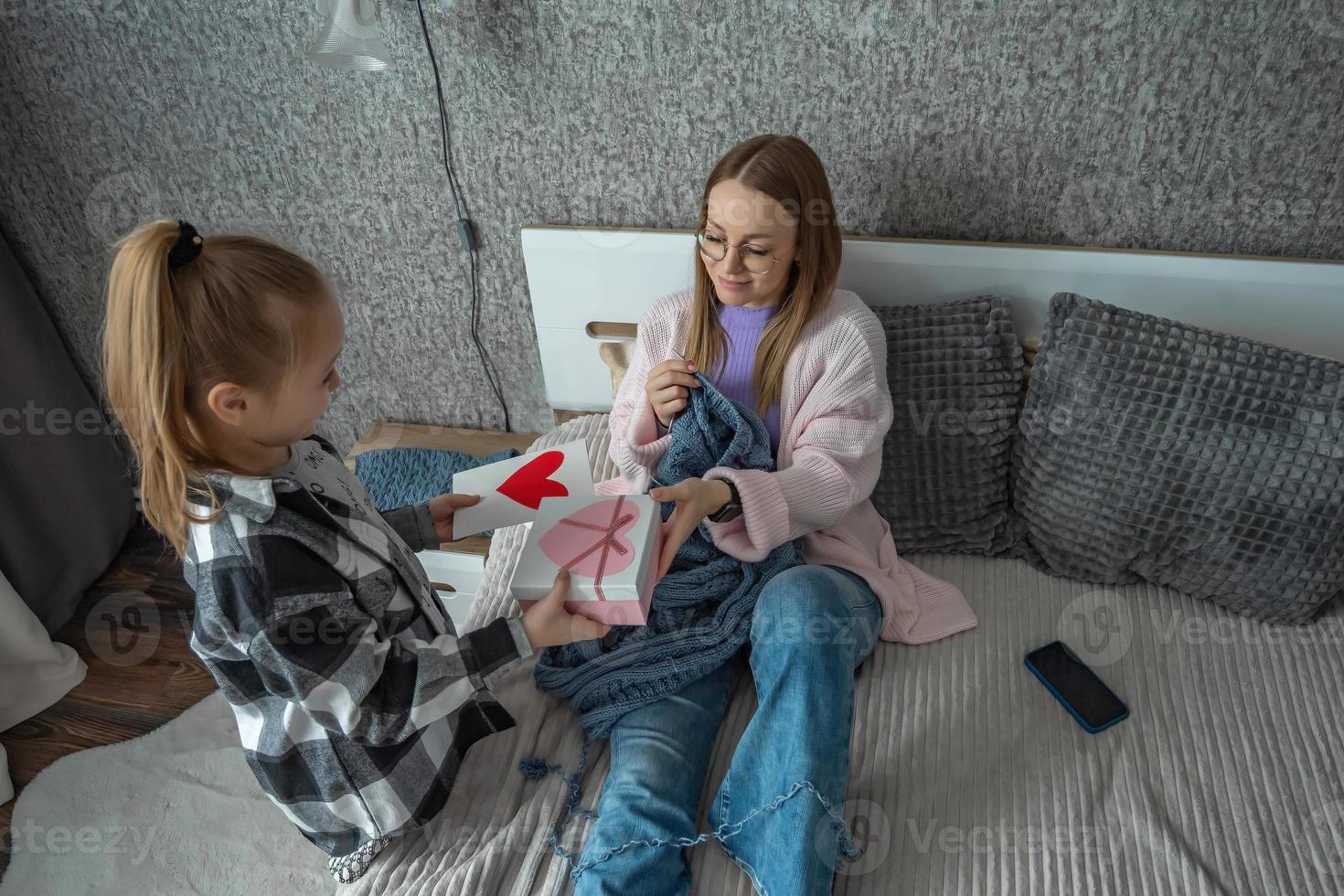 el hija felicita su madre en de la madre día, da su un caja con un regalo y un congratulatorio abrelatas con un corazón hecho por sí misma. un mujer tejer en su dormitorio. foto