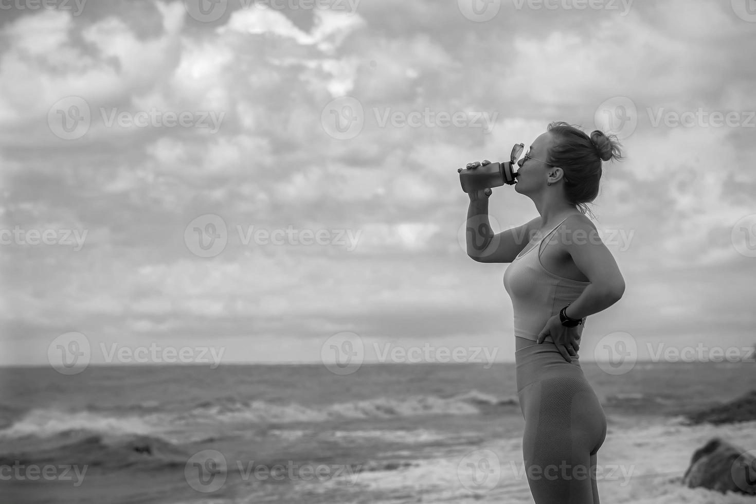 a young beautiful sportswoman in a tracksuit during fitness stands against the background of a sky with clouds, in her hands is a shaker with water that she drinks, copy space, black and white photo