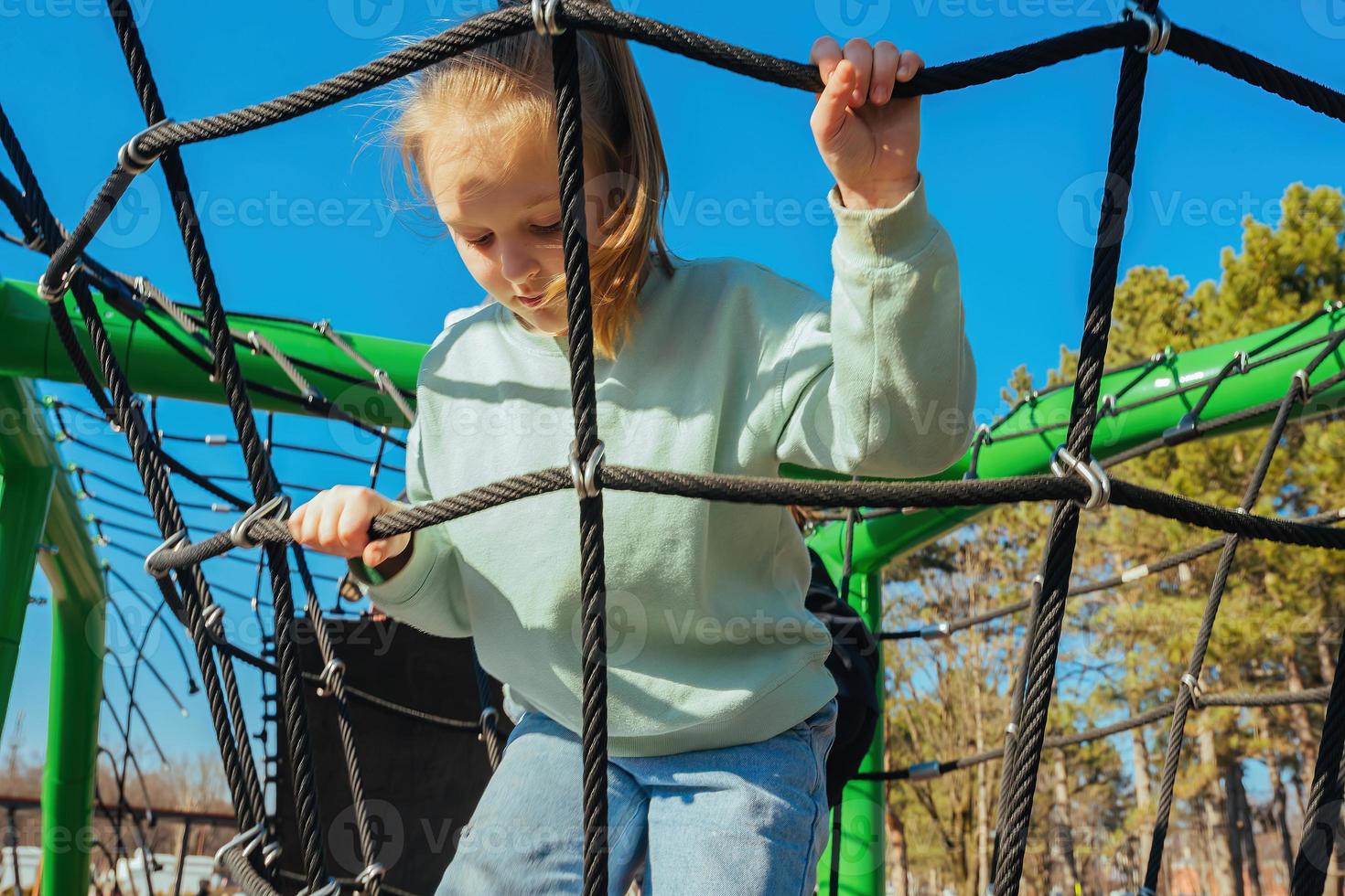 A happy active ten year old girl climbed a rope web on a playground against a bright blue sky on a sunny day photo