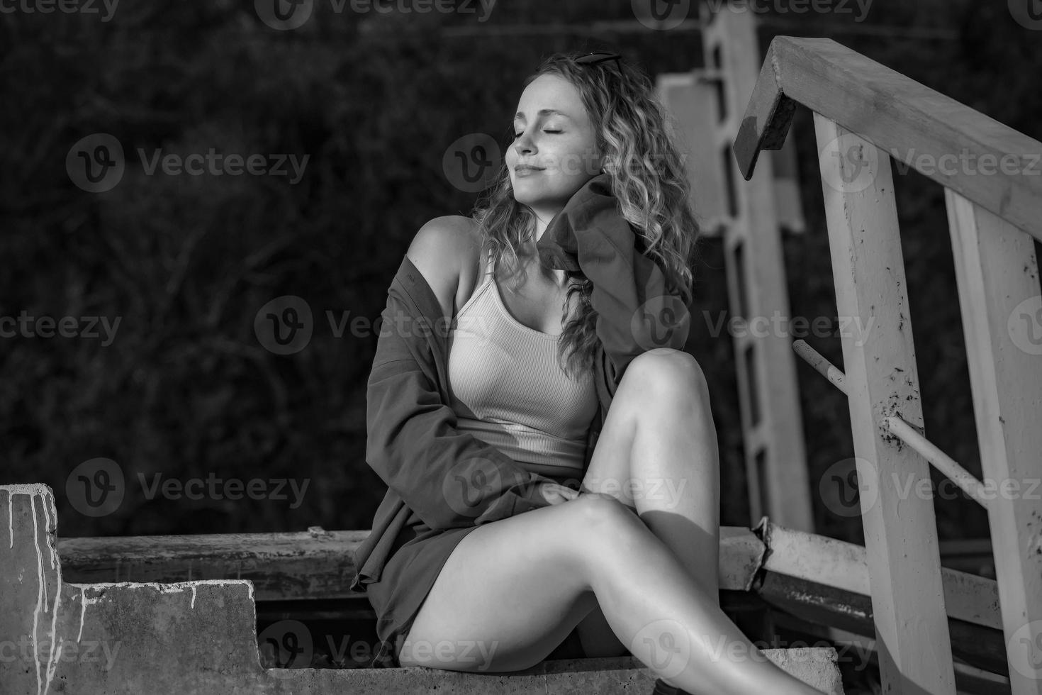 A young beautiful woman with wavy hair is sitting on the stairs leading to the railway in the rays of the setting sun, black and white photo