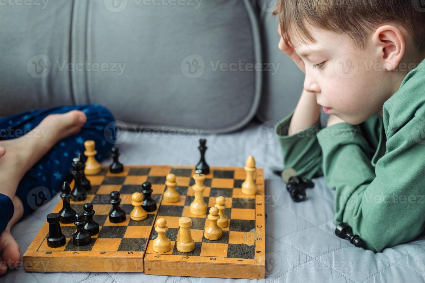 Boys play wooden chess lying on a gray sofa. photo