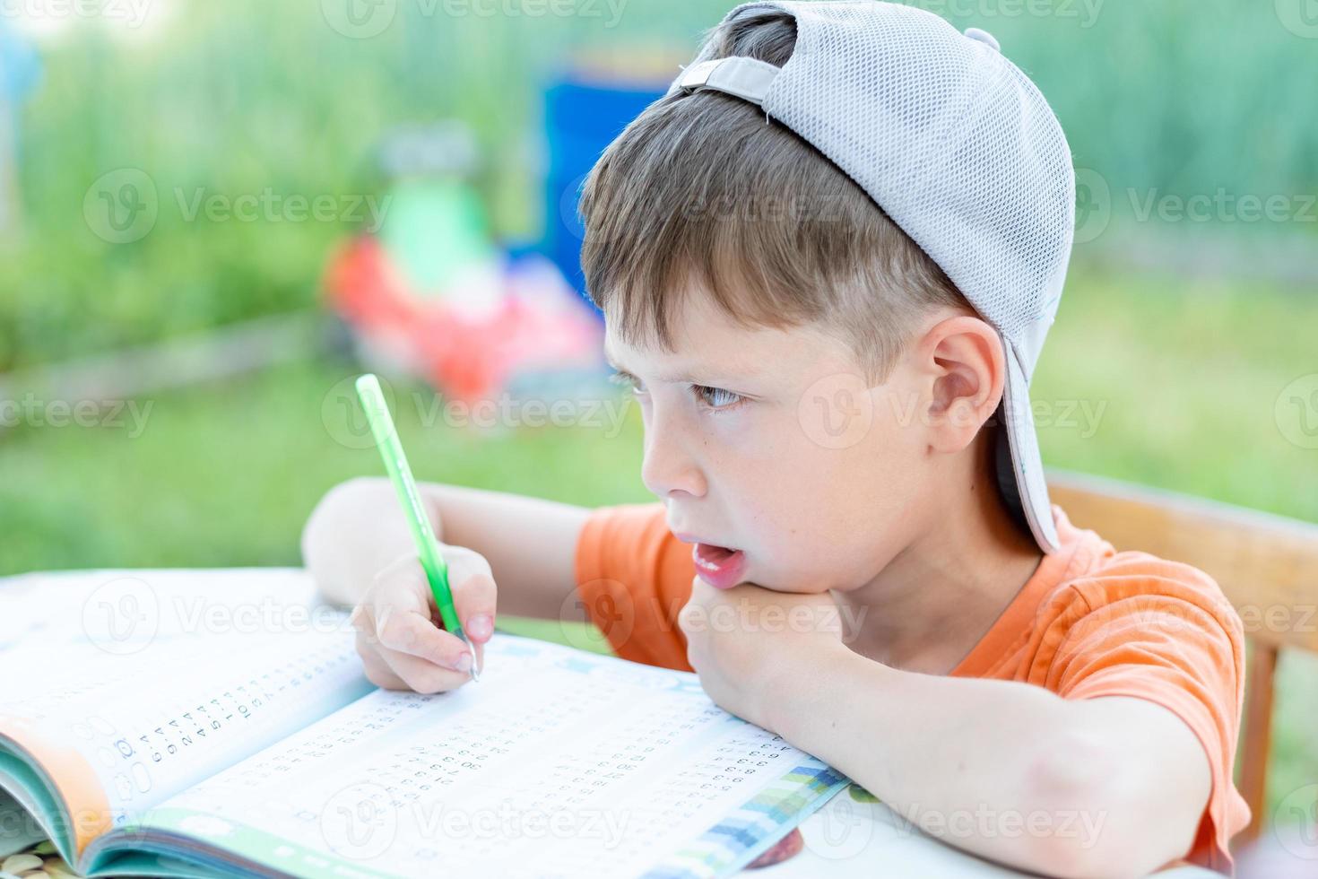 a boy in a cap solves math examples at a table on the street. Child doing homework in the park photo