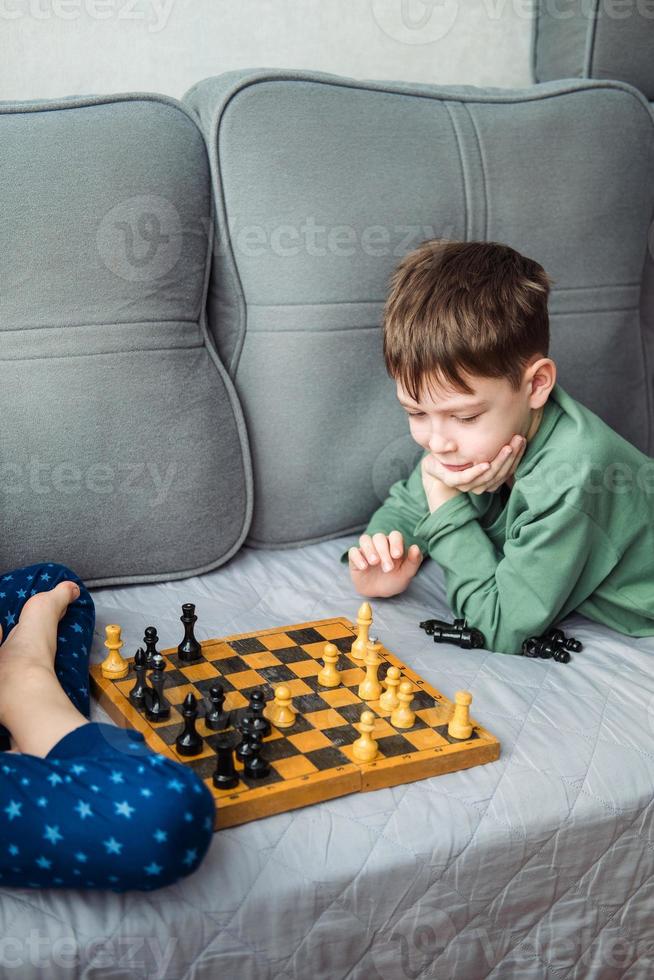 Boys play wooden chess lying on a gray sofa. photo