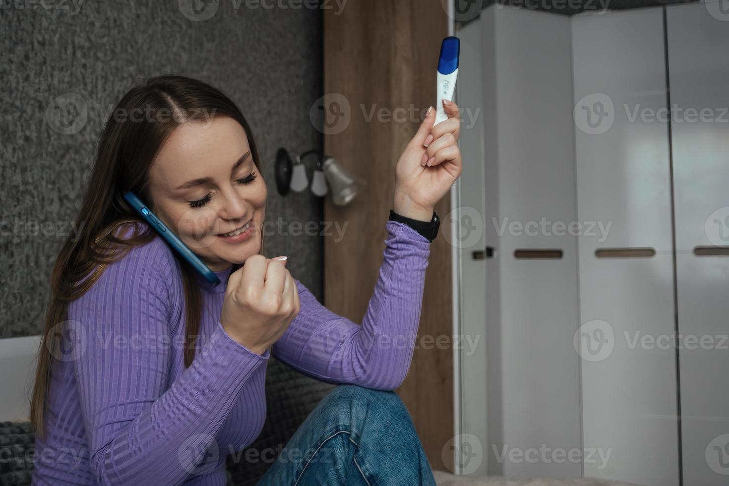 a young beautiful woman rejoices and is happy, in her hands a positive pregnancy test, she shares her joy on the phone photo