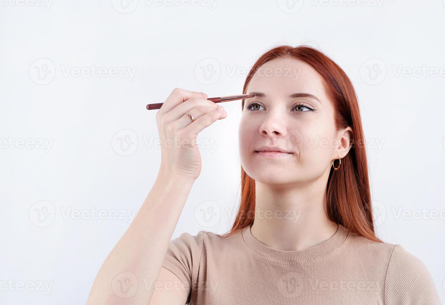 alegre pelirrojo joven mujer utilizando maquillaje cepillo haciendo arriba aislado en blanco antecedentes foto