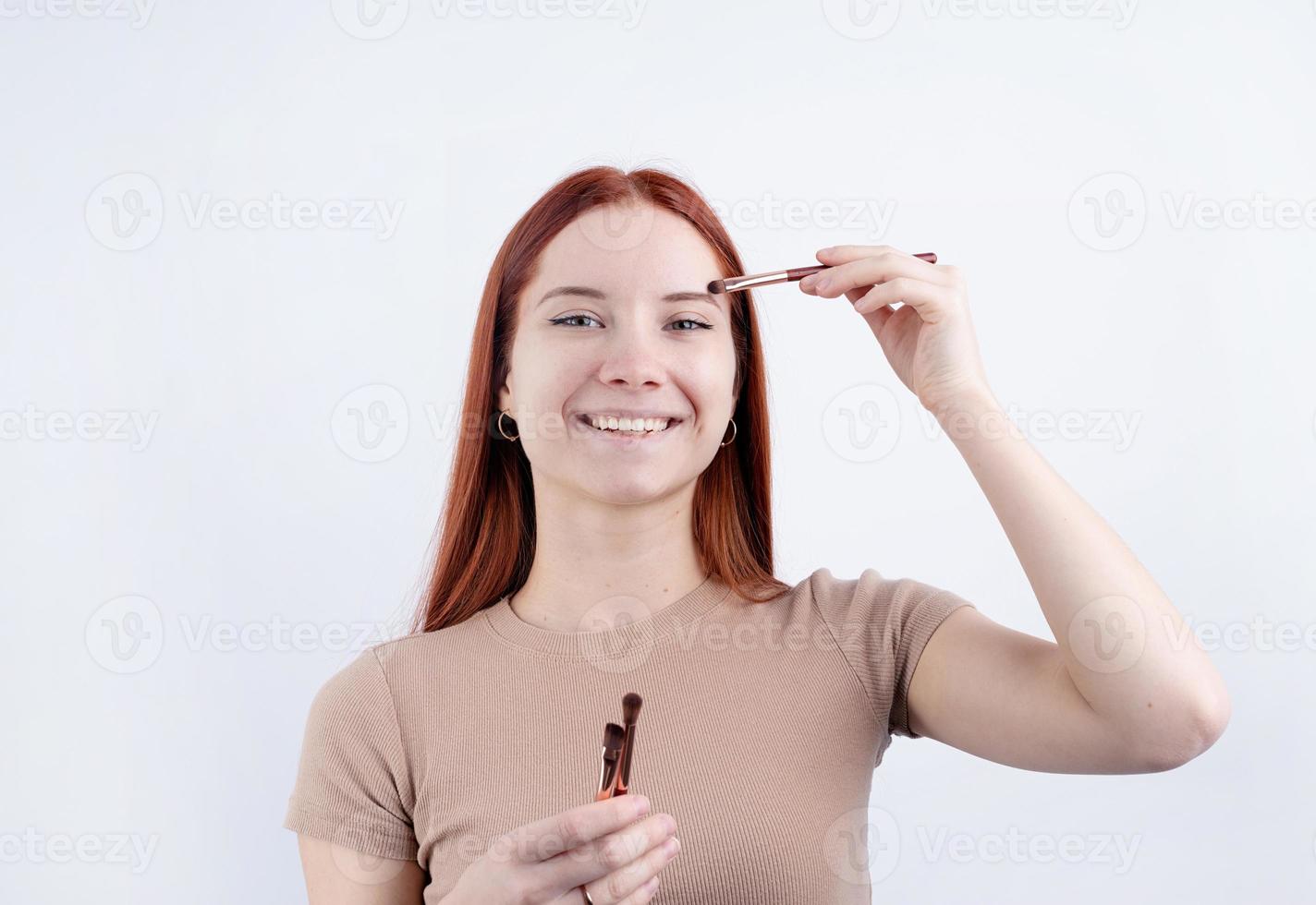 alegre pelirrojo joven mujer utilizando maquillaje cepillo haciendo arriba aislado en blanco antecedentes foto