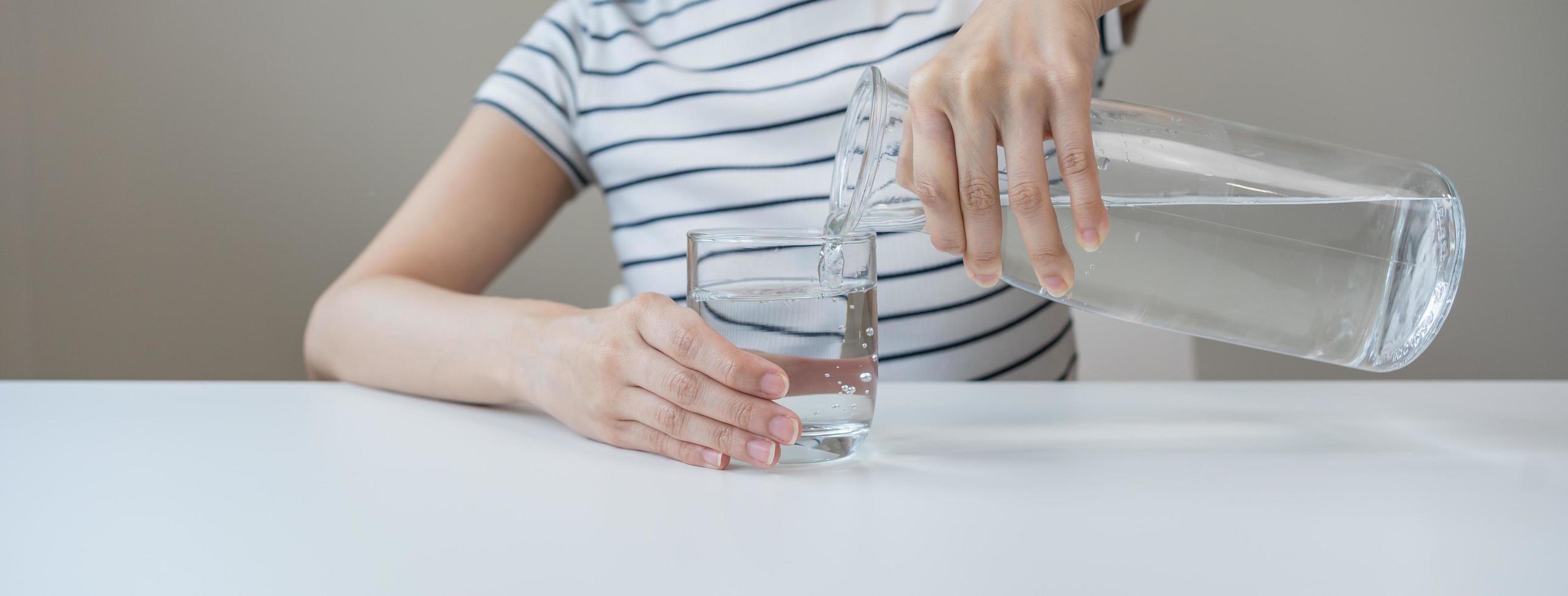 Happy beautiful, thirsty asian young woman, girl drinking, hand in holding, pouring water from jug into transparent glass from pitcher for hydration of body. Health care, healthy lifestyle concept. photo