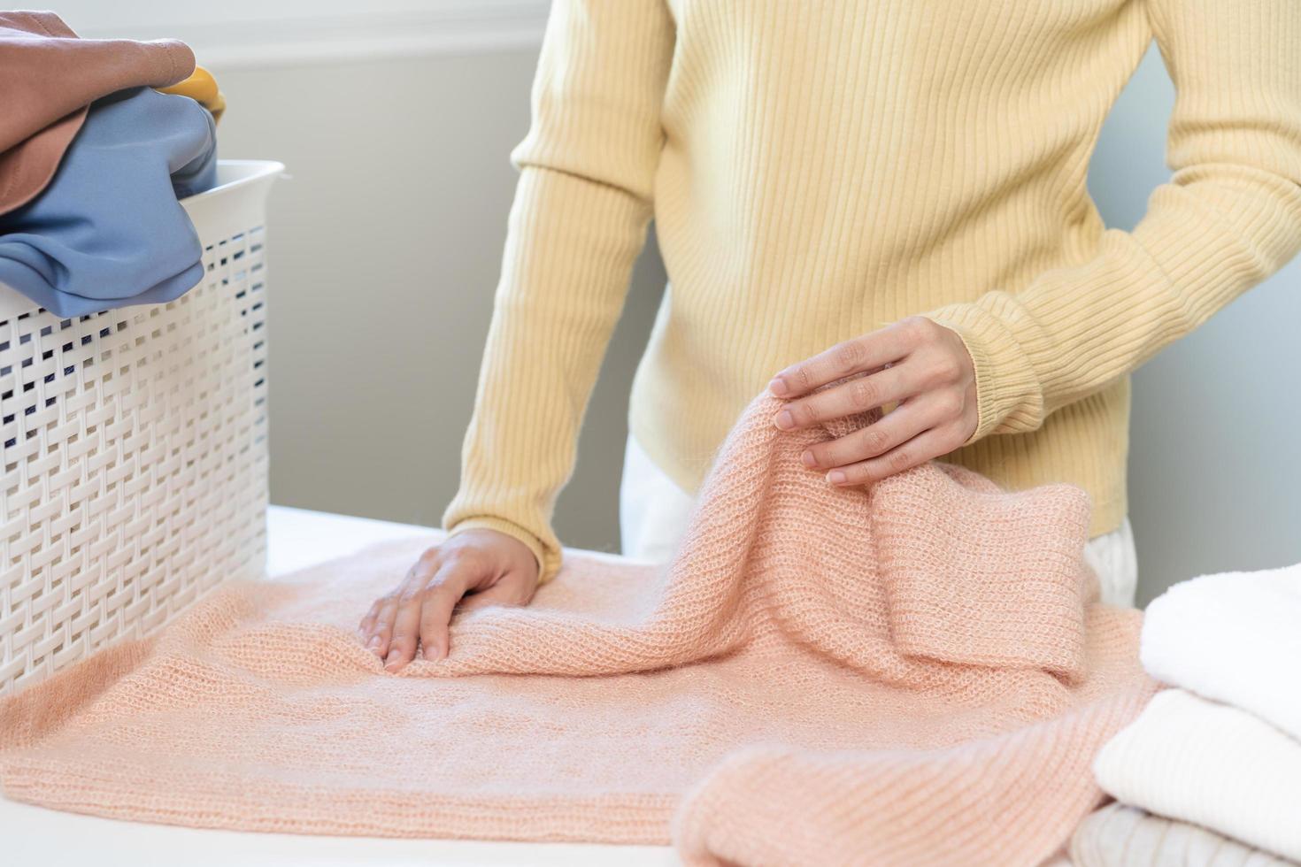 ama de casa, asiático joven mujer mano en muchos plegable recién camisas, suéteres o vestir en escritorio, mesa después Lavado limpiar ropa y el secado, haciendo casa trabajando en habitación a hogar. lavandería y mucama. foto