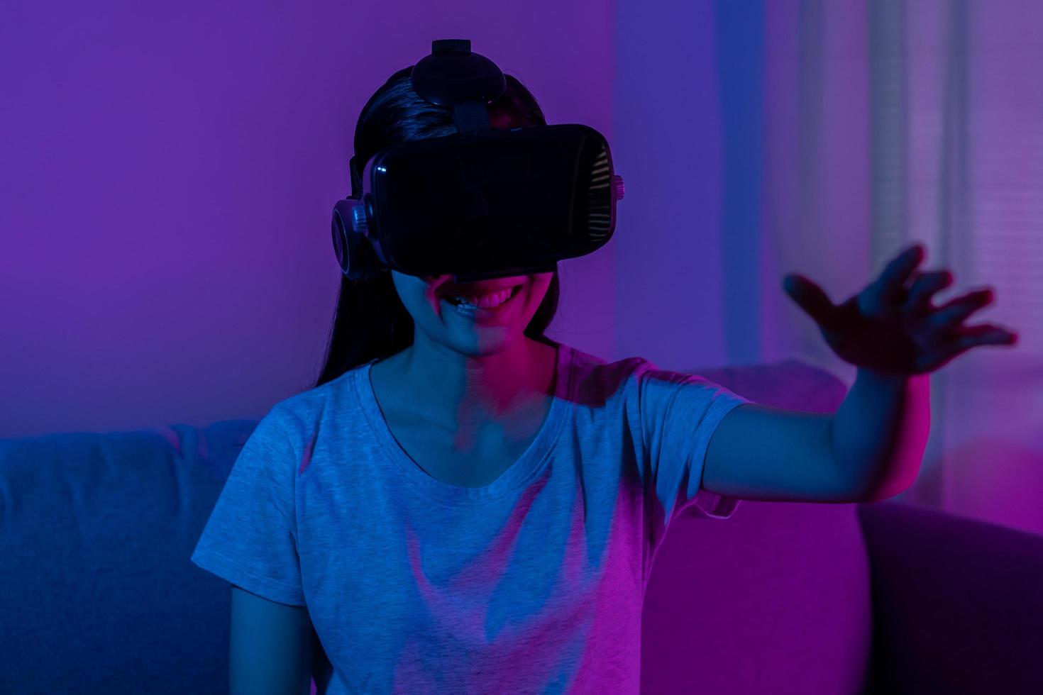 recreación, asiático joven mujer vistiendo vr auriculares o visual realidad gafas de protección, lentes o artilugio, jugar vídeo juego de simulador futuro, futurista mientras usable, sentado en sofá, sofá. foto