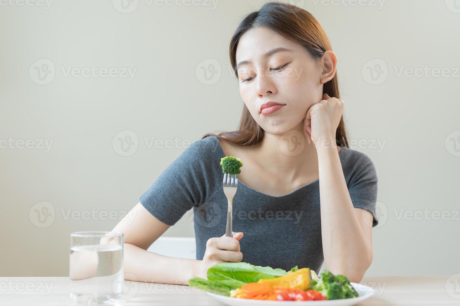 dieta en aburrido, infeliz hermosa asiático joven mujer, niña en haciendo dieta, participación tenedor a brócoli en ensalada lámina, disgusto o cansado con comer Fresco vegetales. nutrición de limpio, sano comida bueno gusto. foto