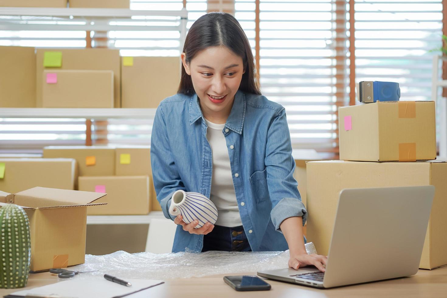 Small business entrepreneur SME, asian young woman owner packing product, checking parcel box delivery, using laptop receive order from customer, working at home office. Merchant online, ecommerce. photo