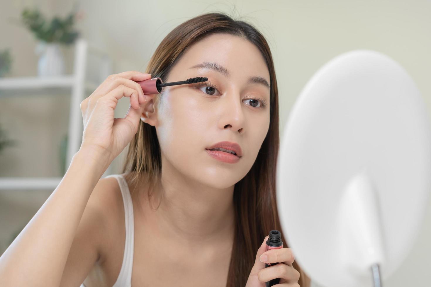 Happy beauty concept, cute asian young woman, girl smile, make up face hand by applying mascara brush on eyelash, looking into mirror in morning routine at home. People look with natural fashion style photo