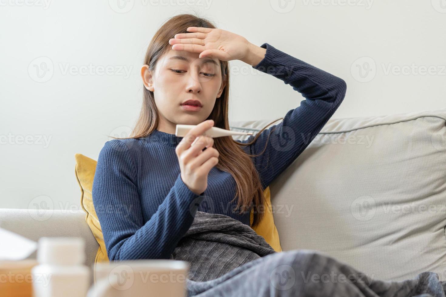 enfermo, influenza asiático joven mujer, niña dolor de cabeza tener un fiebre, gripe y cheque termómetro medida cuerpo temperatura, sensación enfermedad sentado en sofá cama a hogar. salud cuidado persona en virus, COVID-19. foto