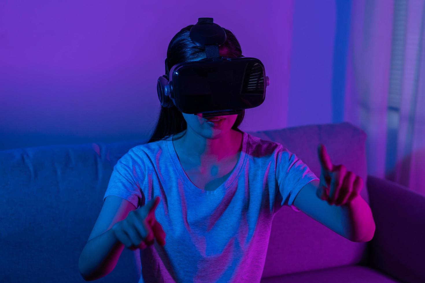 recreación, asiático joven mujer vistiendo vr auriculares o visual realidad gafas de protección, lentes o artilugio, jugar vídeo juego de simulador futuro, futurista mientras usable, sentado en sofá, sofá. foto