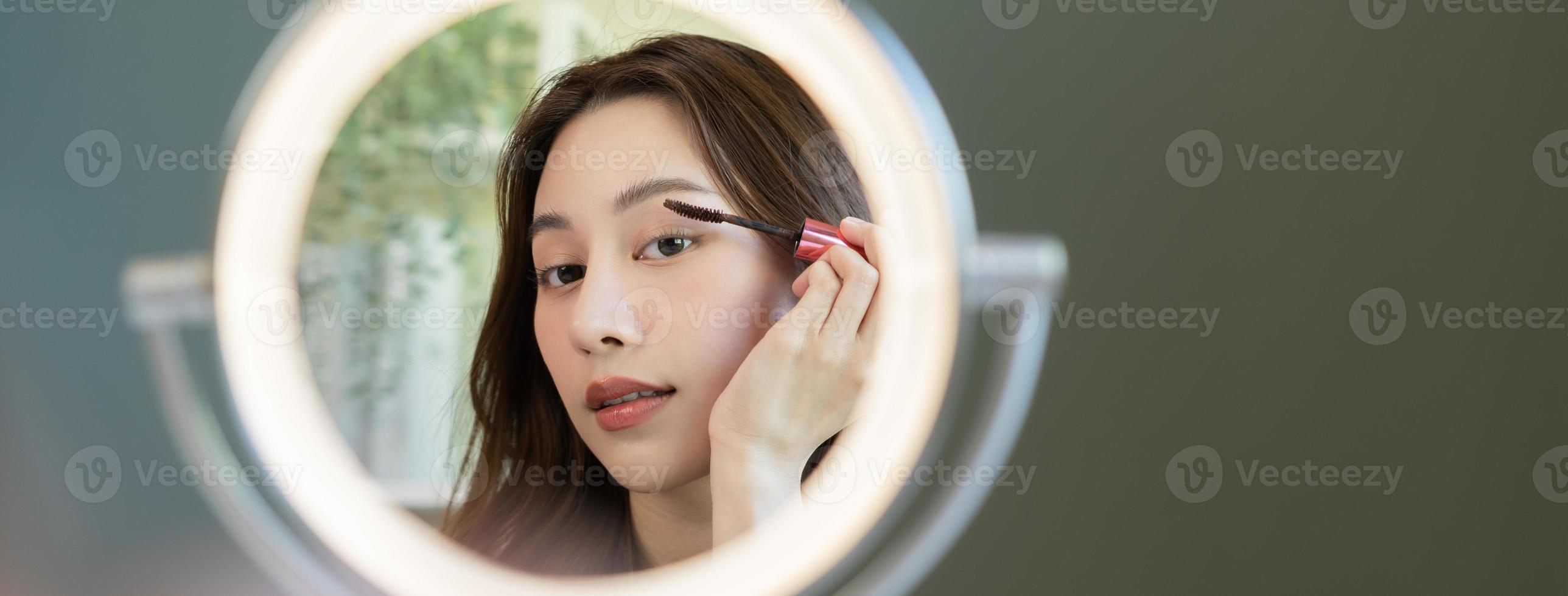 belleza blogger concepto, linda asiático joven mujer, niña hacer arriba cara por aplicando máscara cepillo en ceja, mirando a el espejo a hogar. hembra Mira con natural Moda estilo. foto
