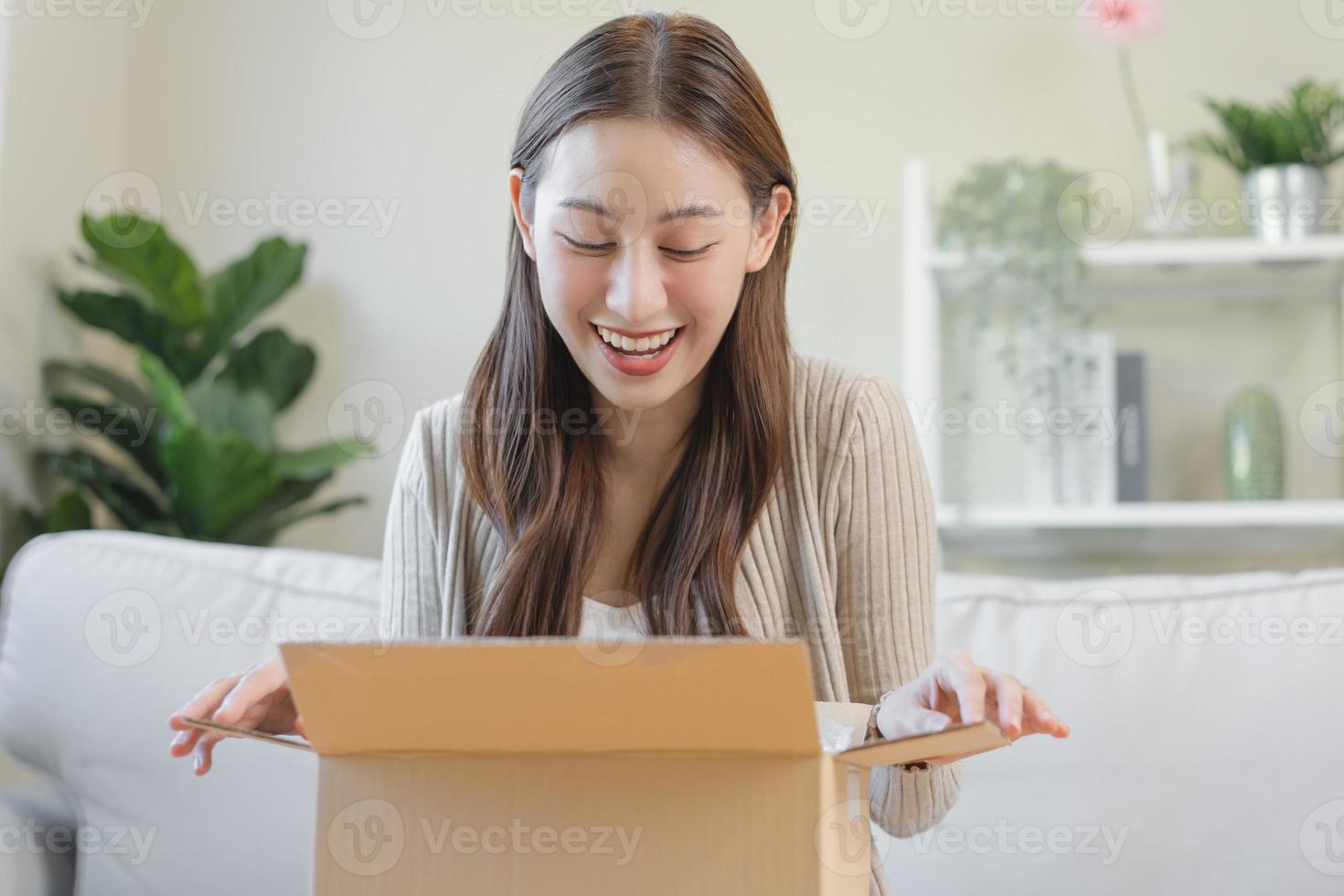 Happy excited, asian young woman, girl customer sitting on sofa at home, opening and unpacking cardboard box carton parcel after buying ordering present, shopping online, delivery service concept. photo