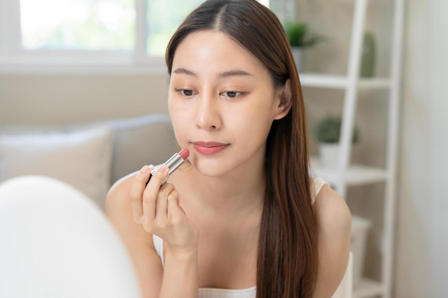 Skin care cosmetics concept, beautiful asian young woman, girl make up face by applying lipstick, lips balm on her mouth, looking at the mirror at home. Female look with natural fashion style. photo