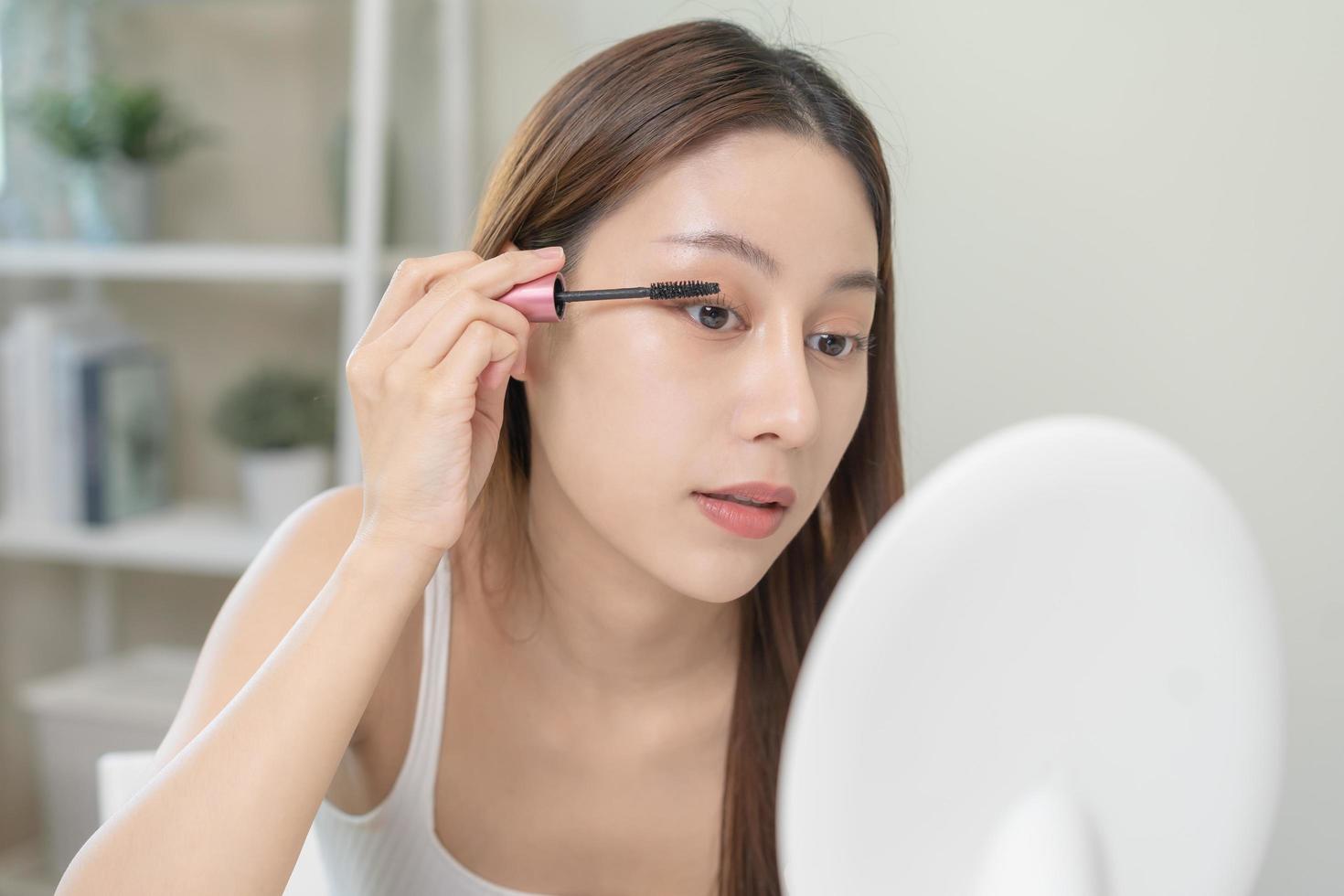 Happy beauty concept, cute asian young woman, girl smile, make up face hand by applying mascara brush on eyelash, looking into mirror in morning routine at home. People look with natural fashion style photo