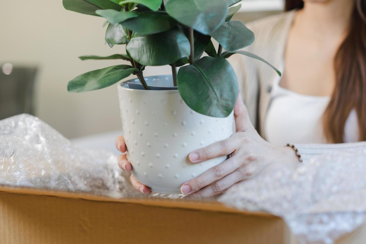 Asian young  customer woman hand in received, unpack package carton open parcel in cardboard box, holding ceramic plant pot. Moving, preparing in new house, relocation. Online shop delivery at home. photo