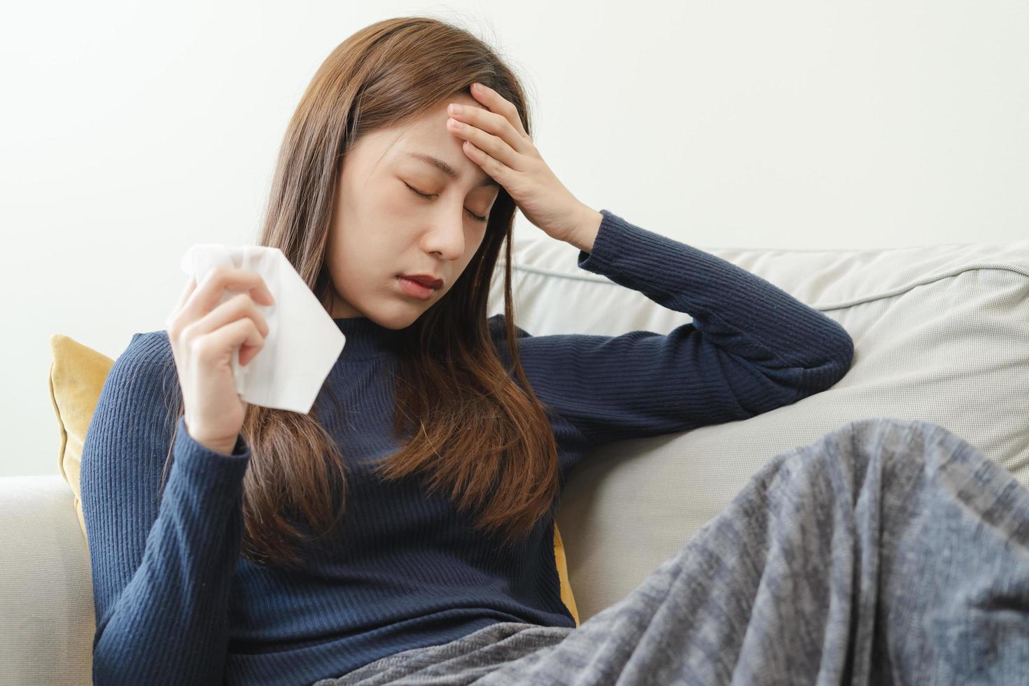 enfermo, influenza asiático joven mujer, niña dolor de cabeza tener un fiebre, gripe y cheque medida cuerpo temperatura, sensación enfermedad sentado en sofá cama a hogar. salud cuidado persona en virus, COVID-19. foto