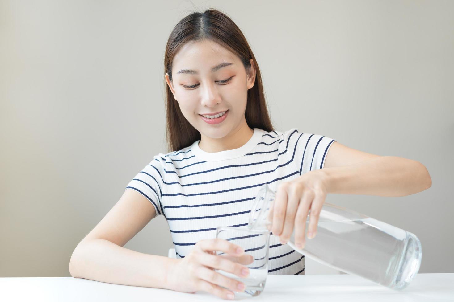 Happy beautiful, thirsty asian young woman, girl drinking, hand in holding, pouring water from jug into transparent glass from pitcher for hydration of body. Health care, healthy lifestyle concept. photo