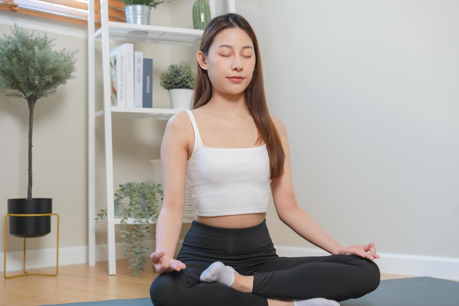 ejercicio, calma asiático joven mujer, niña mano en calma actitud sentado práctica meditando en loto posición en estera a hogar, meditación, ejercicio para bienestar, sano cuidado. relajación, contento ocio. foto