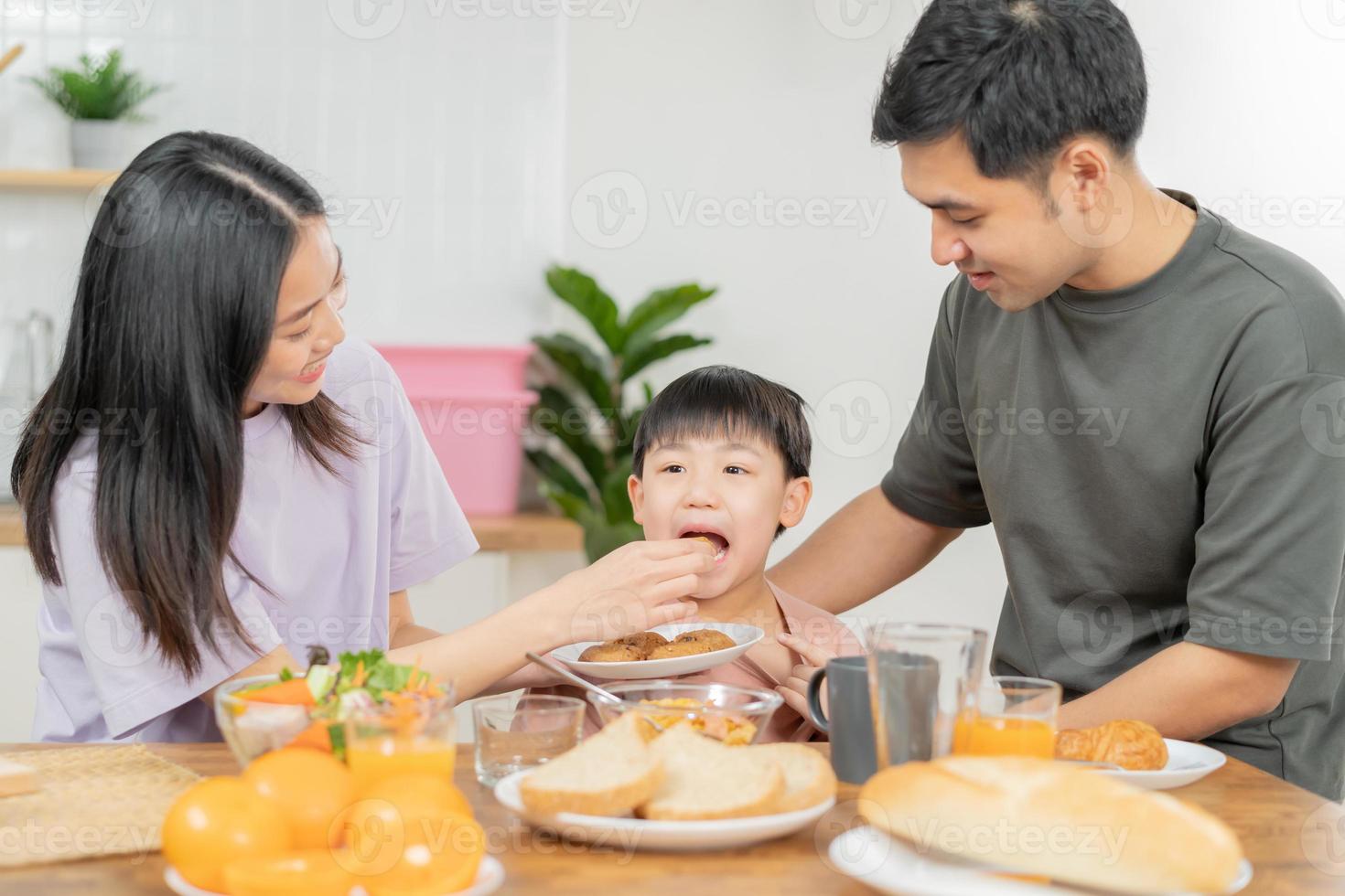 contento refresco familia desayuno en mañana, asiático joven padre padre, madre y pequeño linda chico, niño teniendo comida en cocina comiendo juntos a hogar. alegre, disfrutar Cocinando gente. foto