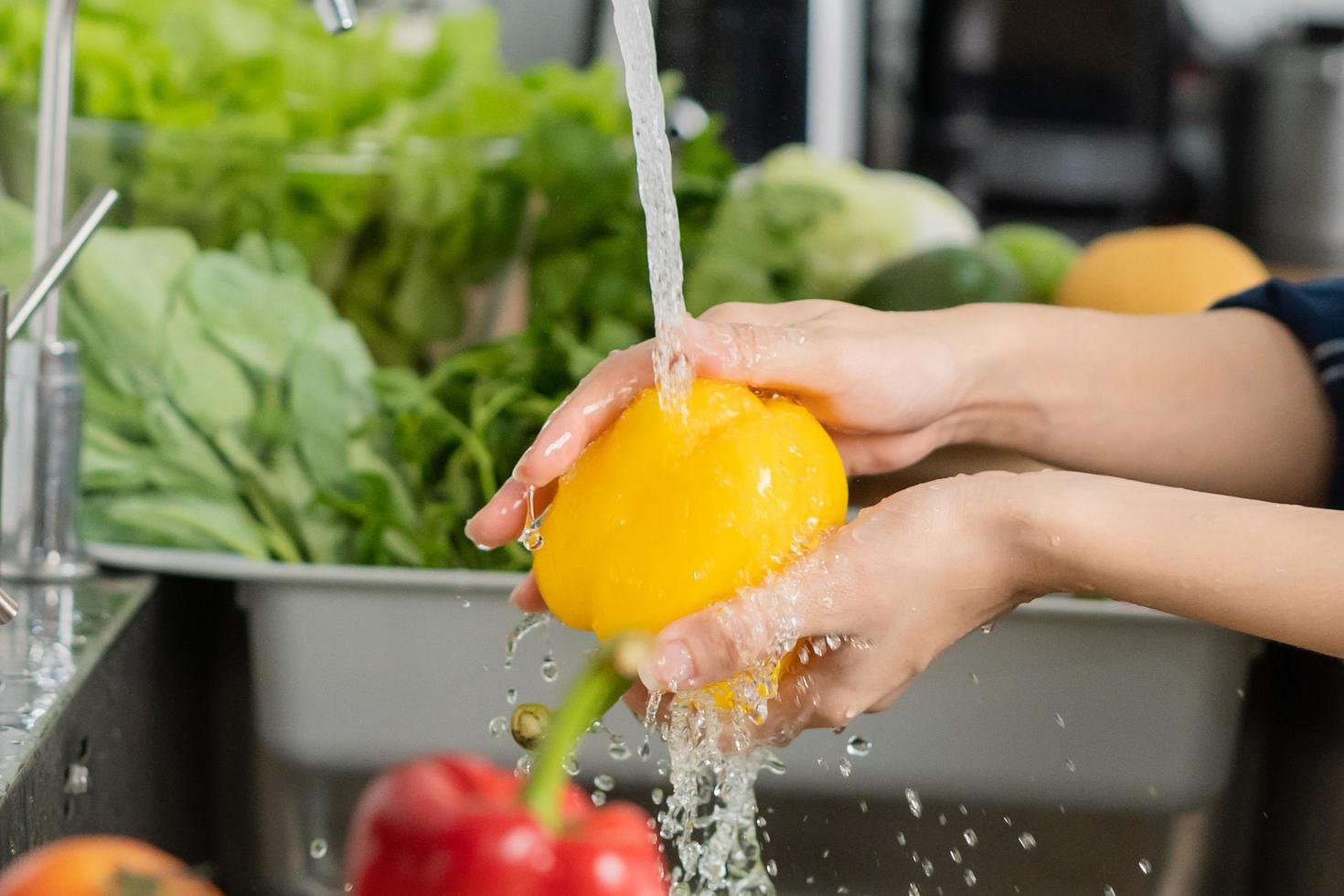 cerca arriba asiático joven ama de casa mujer, Lavado dulce pimienta, amarillo pimenton, vegetales con chapoteo agua en cuenca en lavabo en cocina, preparando Fresco ensalada, Cocinando comida. sano comida gente. foto