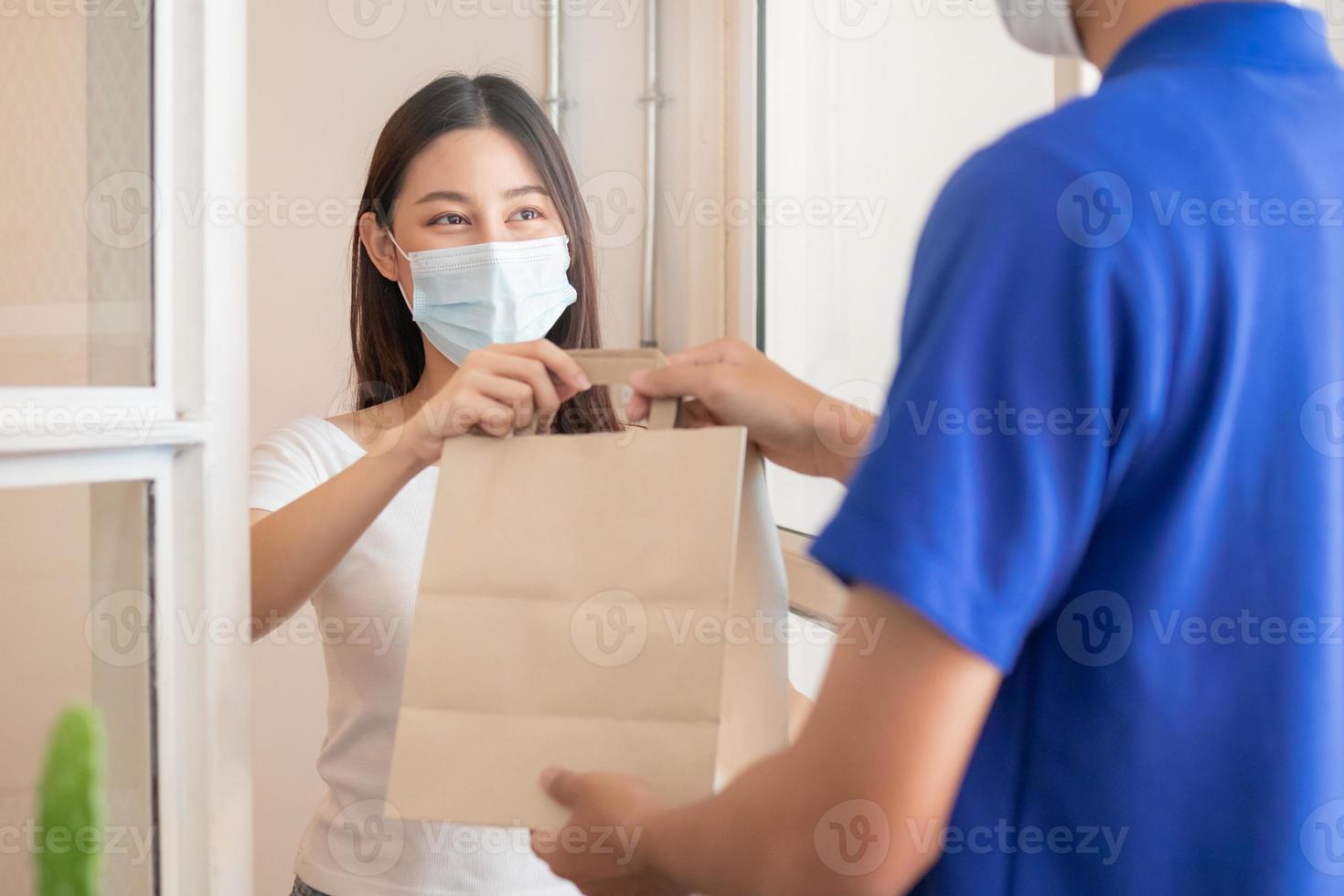 Smile, Happy asian young woman or girl  wearing face mask protect to pandemic of covid-19 and received paper bag from delivery man, messenger in uniform while lockdown, quarantine at front door home. photo