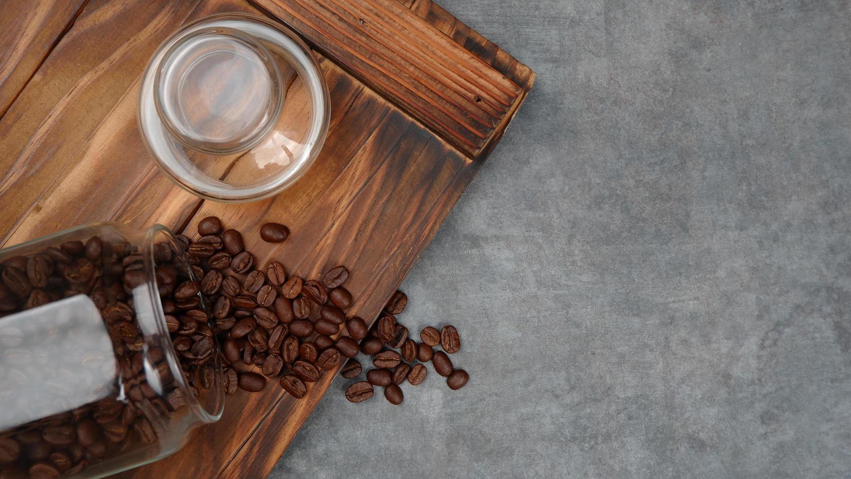 Photography of coffee beans spilling from a jar, on pieces of wood with a gray background, suitable for photos of food and beverage products, with landscape photo format