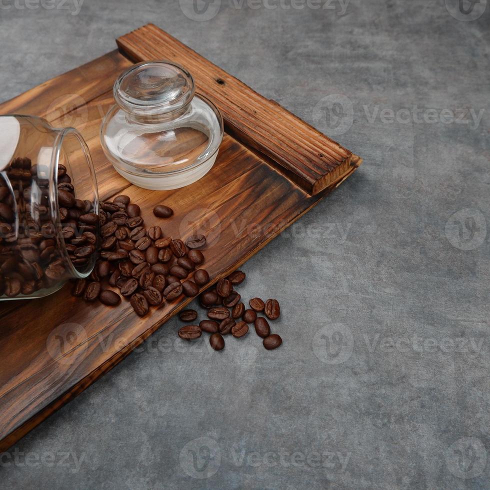 Photography of coffee beans spilling from a jar, on pieces of wood with a gray background, suitable for photos of food and beverage products, with perspective angle square photo format