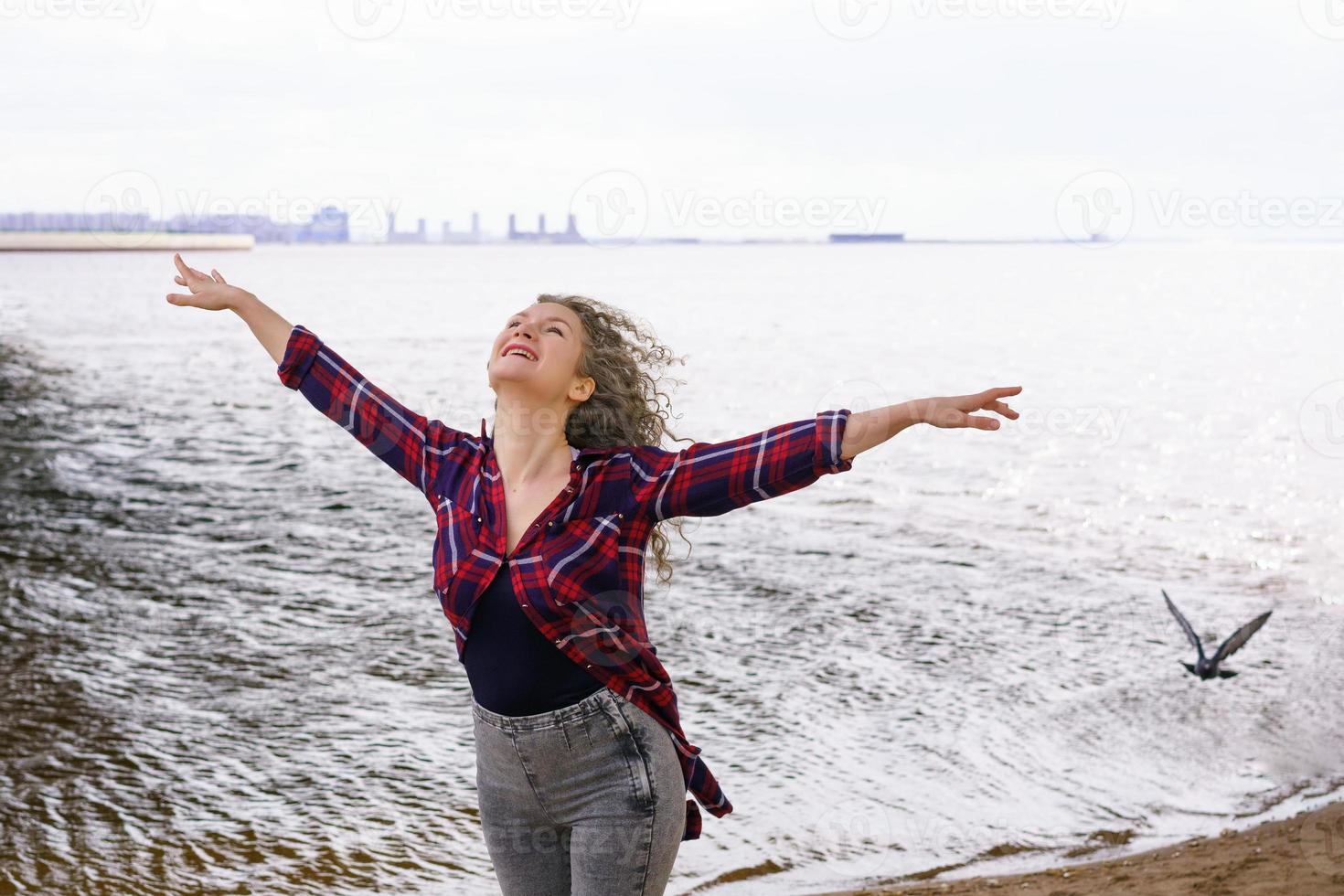 un joven mujer en primavera posando en el río banco, con Rizado pelo se extiende su brazos a el lado, el concepto de libertad foto