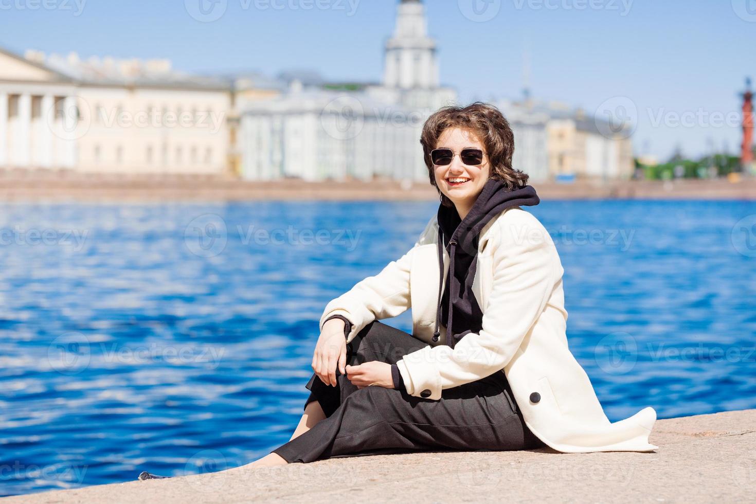 Girl sits on concrete flooring embankment in white coat and black sweatshirt photo