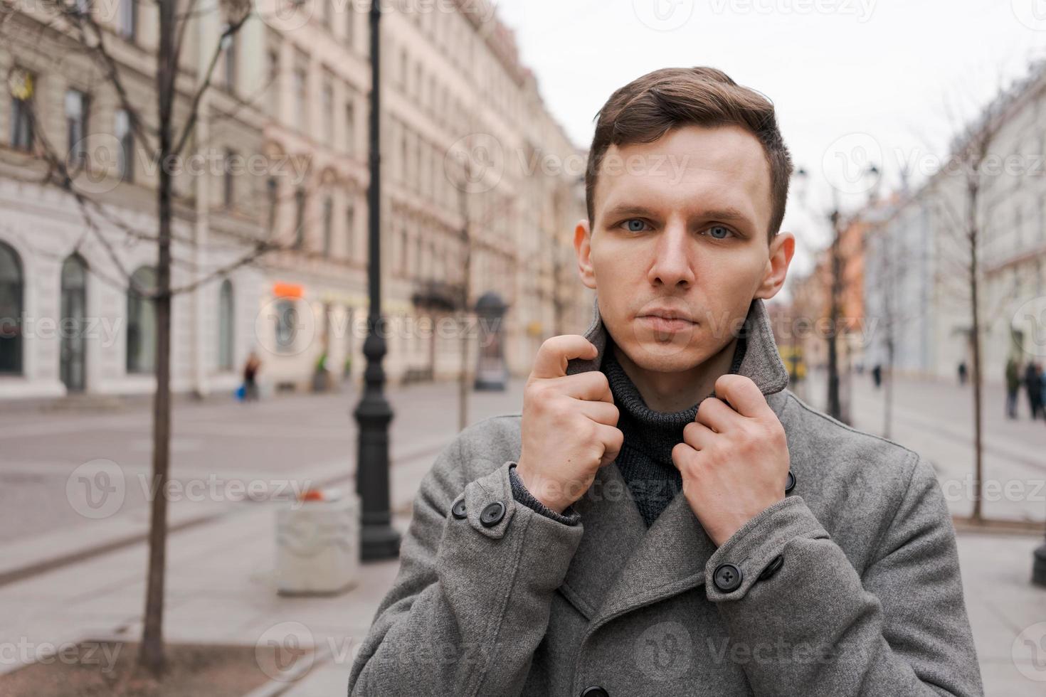 Young handsome man in gray coat walks on the street in city in gray weather photo