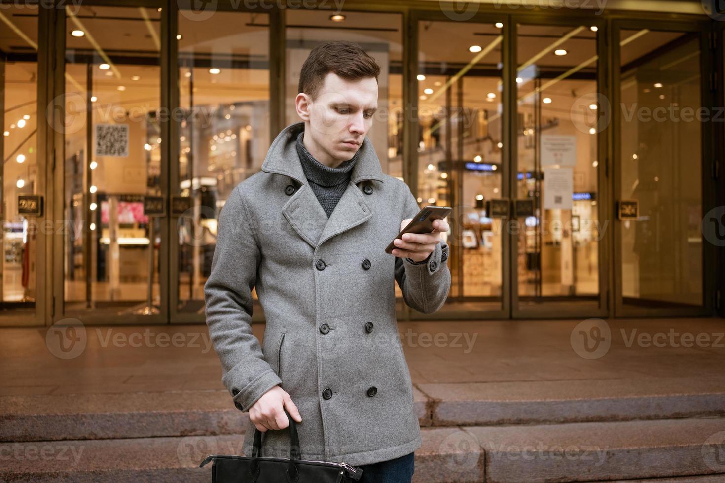 Smiling young man in gray coat is holding scroll text messages in his mobile photo