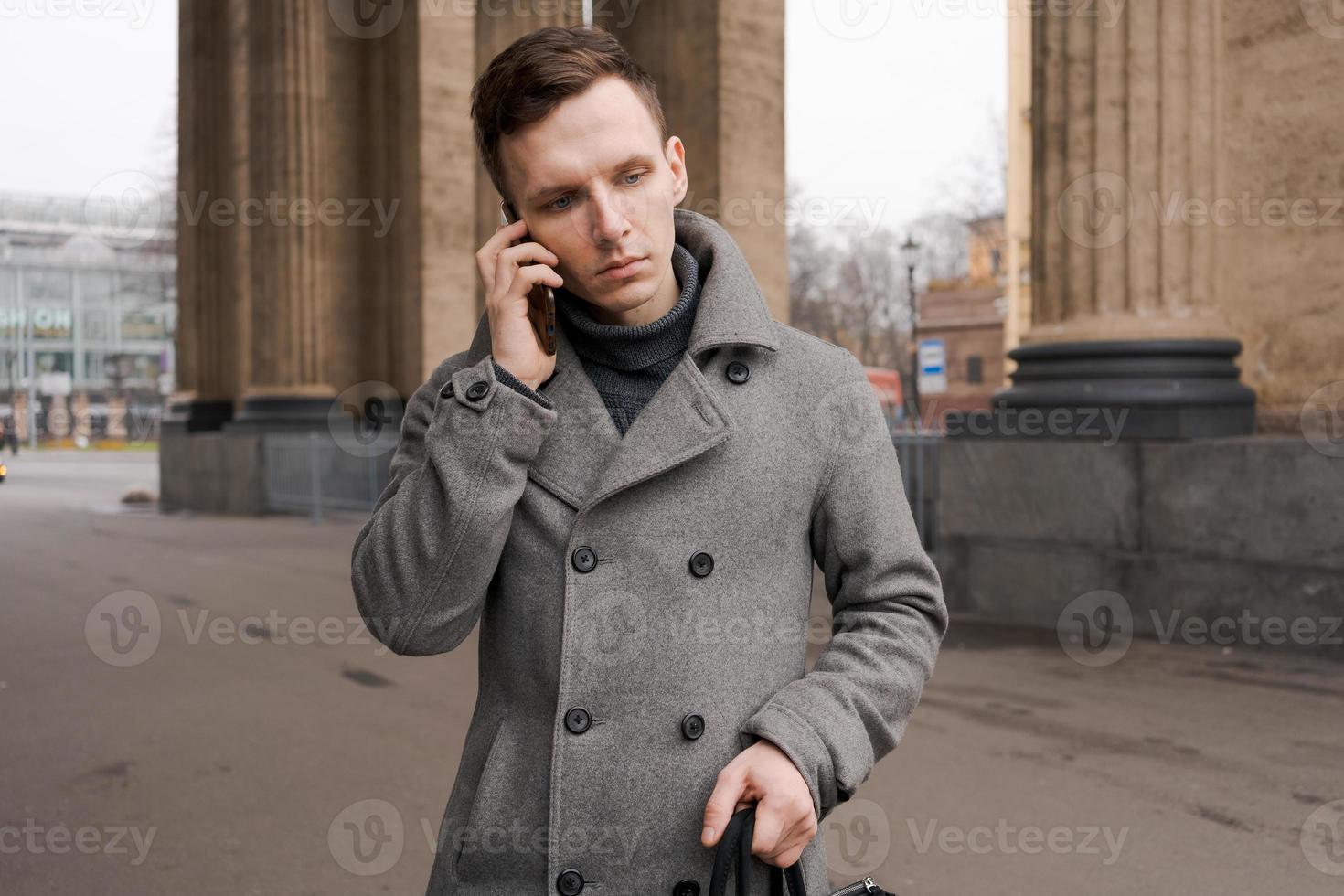 hombre en gris Saco y suéter soportes en ciudad calle. negociaciones en teléfono. resuelve foto