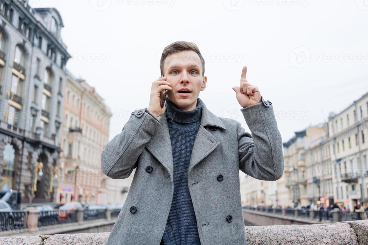 Man in gray coat and sweater stands on city street. Talks on phone. Solves photo