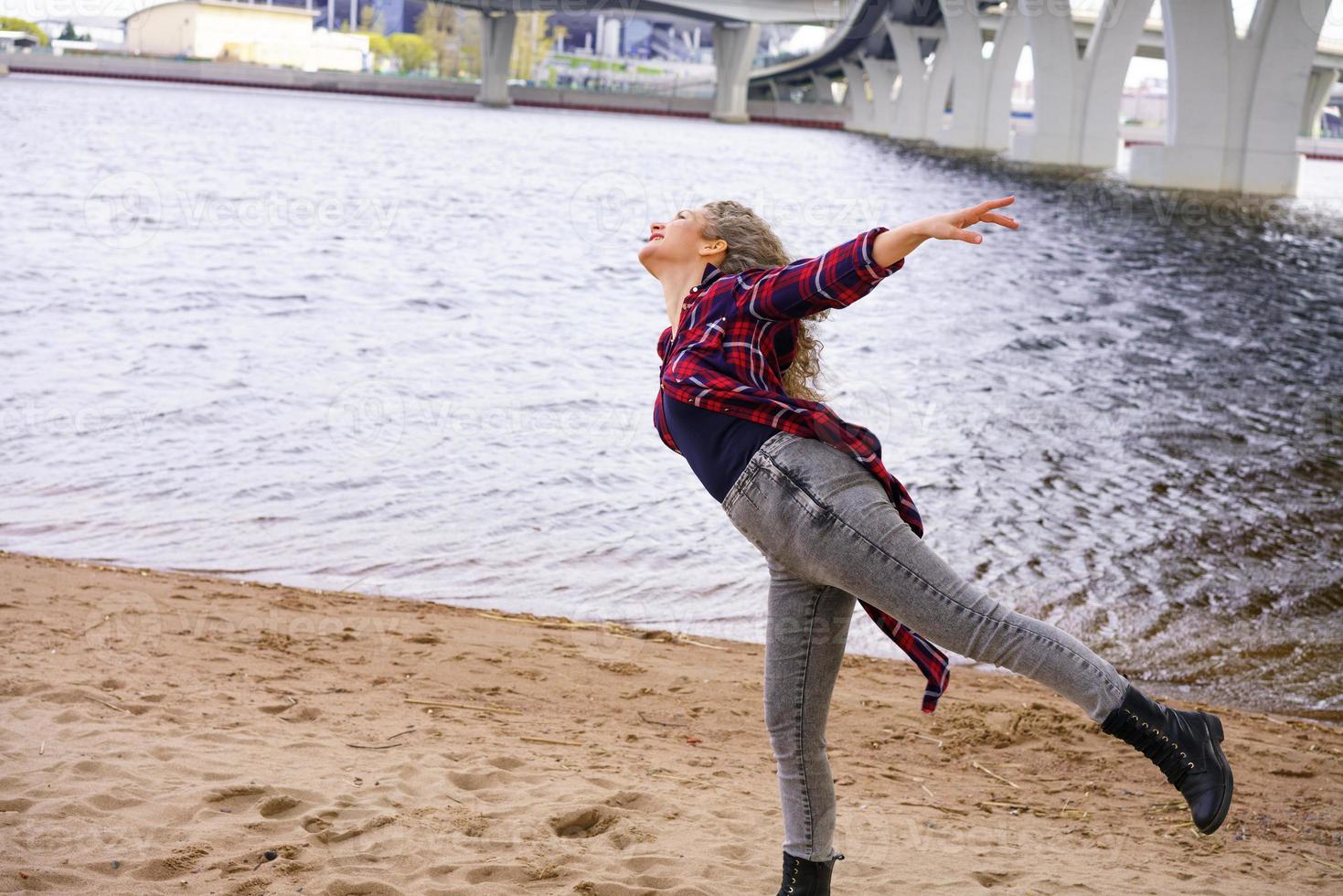 un joven mujer en primavera posando en el río banco, con Rizado pelo se extiende su brazos a el lado, el concepto de libertad foto