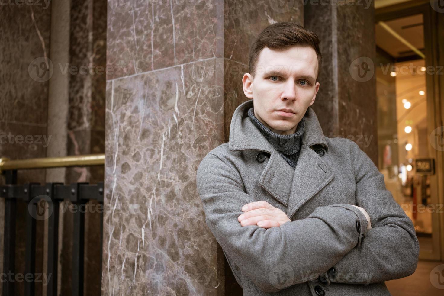 A young man stands outside a shop on the street in a gray coat leaning. photo