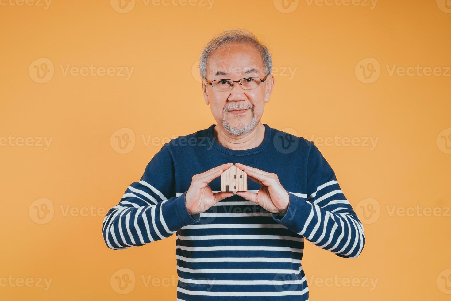 Senior male holding wooden house model property owner after retirement on yellow background. photo